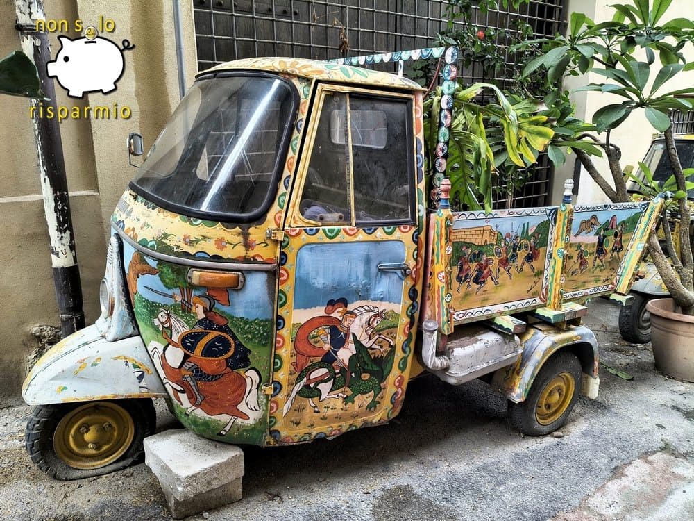 Un'Ape Piaggio d'epoca decorata nello stile tradizionale del carretto siciliano con dipinti folcloristici di cavalieri e battaglie cavalleresche su cabina e cassone, parcheggiata in un vicoletto cittadino