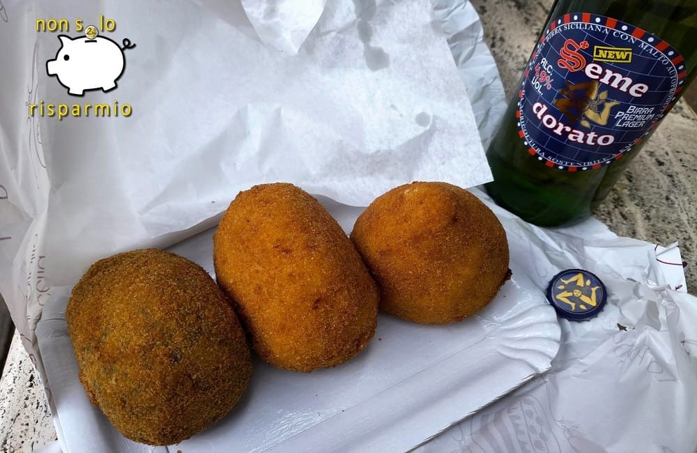 Tre arancine dorate e fritte disposte su un vassoietto di carta, affiancate da una bottiglia di birra artigianale siciliana e dal suo tappo con il simbolo della Trinacria