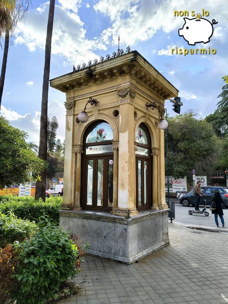 Chiosco d'epoca in stile Liberty in pietra e muratura chiara con finestre ad arco in legno, insegna "Bibliochiosco" e decorazioni in ferro battuto, situato ai margini di un parco cittadino di Palermo