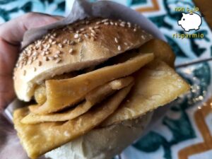 Primo piano di un panino tondo con semi di sesamo, tenuto in mano e farcito con panelle sottili di farina di ceci fritte e crocchè di patate