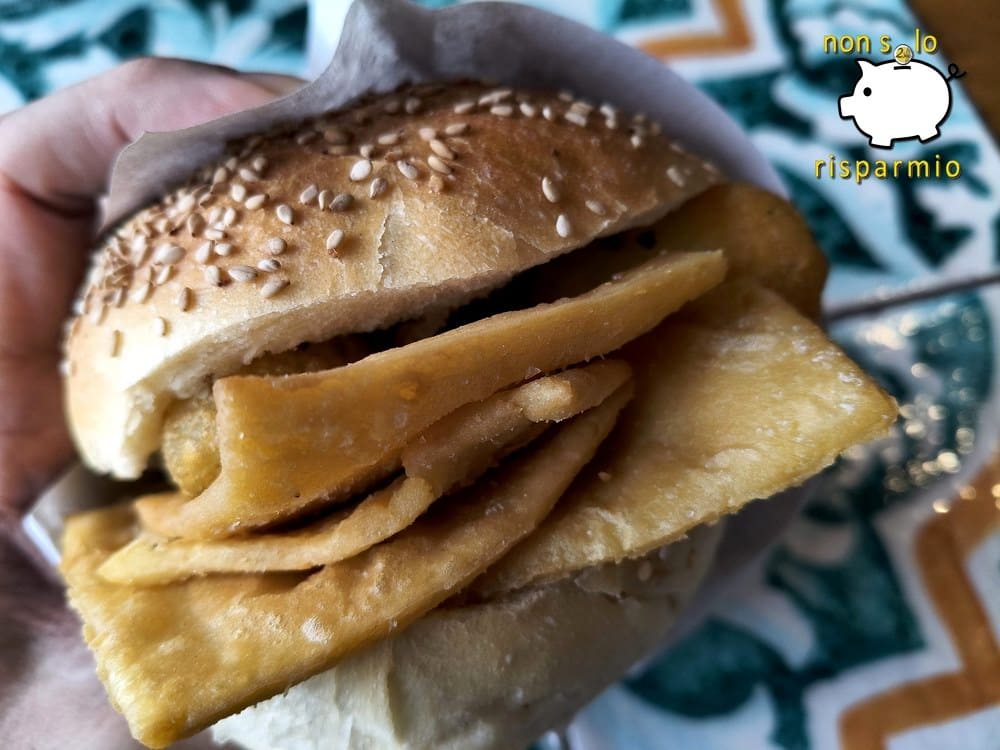 Primo piano di un panino tondo con semi di sesamo, tenuto in mano e farcito con panelle sottili di farina di ceci fritte e crocchè di patate