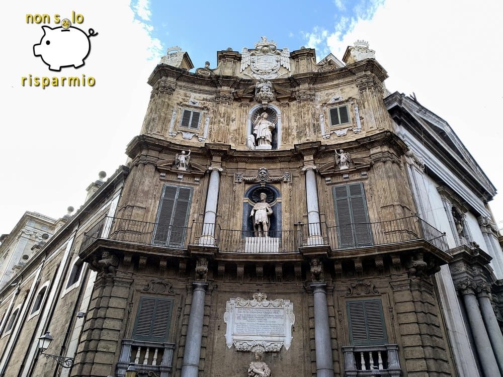 Vista prospettica dal basso della facciata barocca dei Quattro Canti a Palermo affiancata da un edificio adiacente, sotto un cielo azzurro con nuvole.